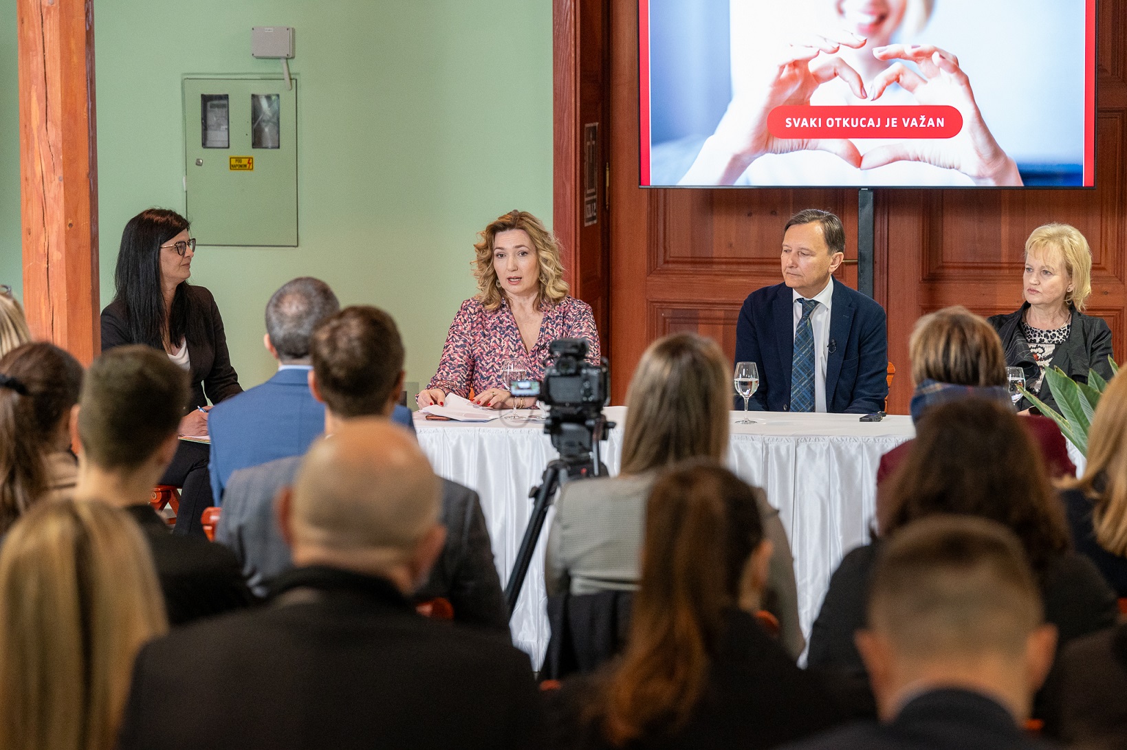 Na fotografiji je prikazana konferencija ili sastanak. Četiri osobe sjede za stolom, a lica su im zamagljena. Publika je okrenuta prema osobama za stolom, a u prvom planu je kamera koja snima događaj. Prostorija ima zelene zidove i drvene elemente, što joj daje elegantan izgled. S lijeve strane zida iza osoba koje sjede nalazi se izlazna vrata.