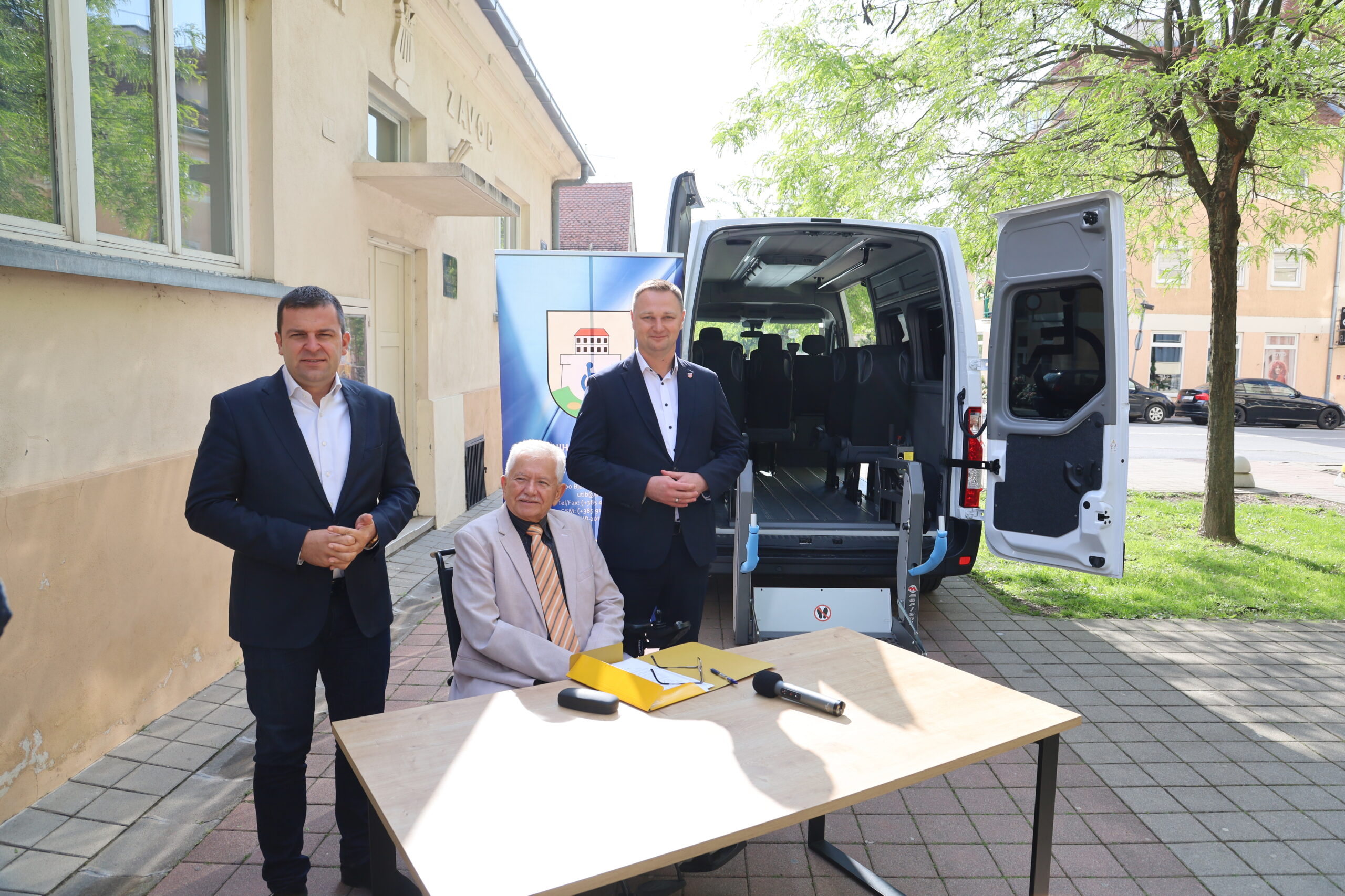 Na fotografiji se nalaze tri osobe odjevene u formalnu odjeću koje stoje ispred stola. Na stolu je mikrofon i nekoliko dokumenata. U pozadini se vidi otvoreni kombi, zgrada i drveće. Izgleda kao da se priprema neka vanjska profesionalna aktivnost ili događaj.