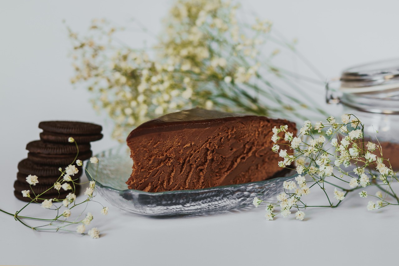 Na slici je komad čokoladne torte na staklenom tanjuru, okružen cvjetnim aranžmanom i čokoladnim keksima s kremom. Pozadina je svijetla i jednostavna, što ističe tamne tonove torte i keksa.