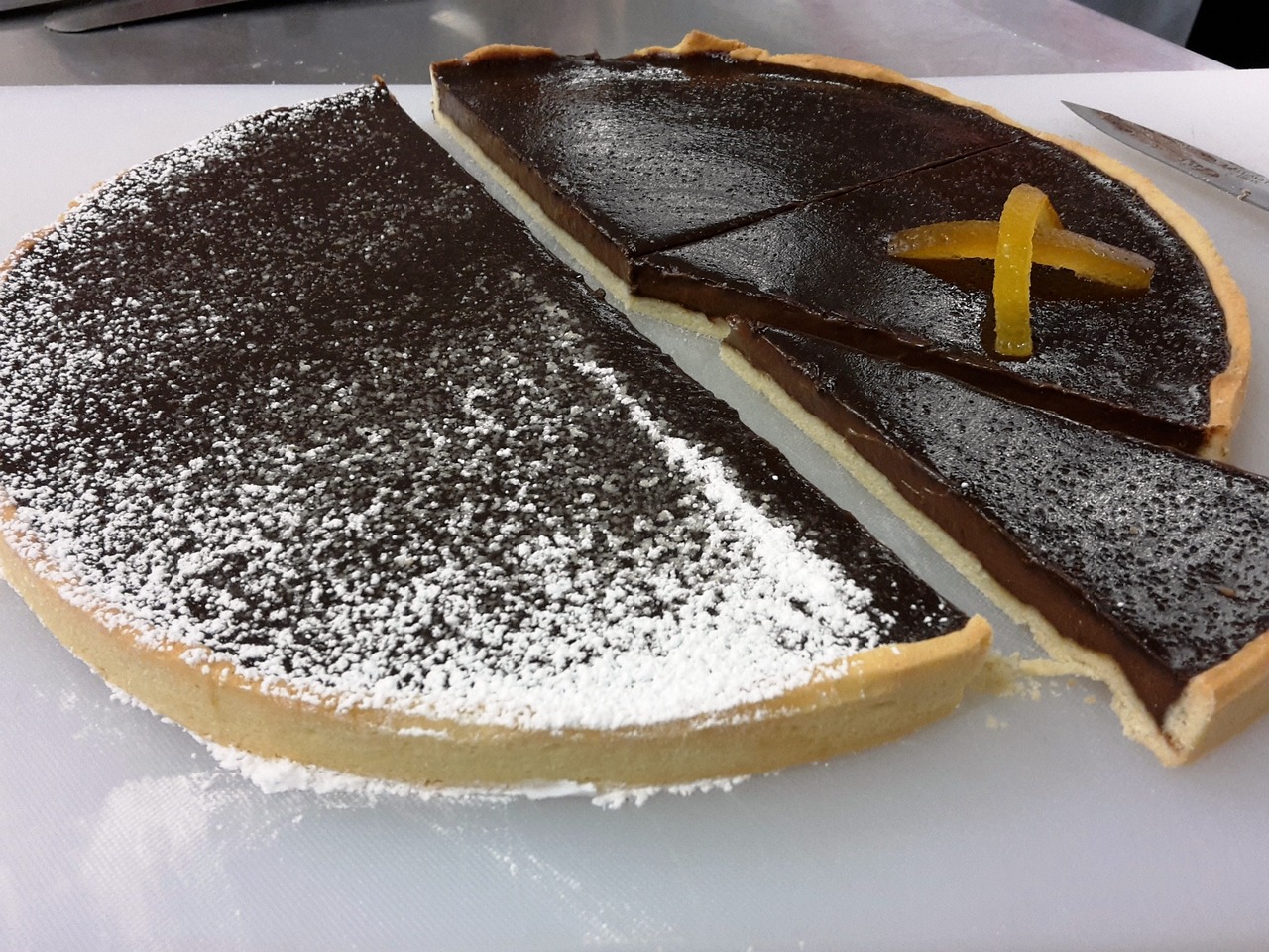 Fotografija prikazuje veliki okrugli tart prekriven tamnom čokoladnom glazurom. Na jednoj polovici tarta posipan je šećer u prahu, stvarajući kontrast u boji i teksturi. Druga polovica tarta ukrašena je križem napravljenim od kriški naranče, dodajući živopisan detalj tamnoj čokoladi. Tart izgleda svježe pripremljen i postavljen je na bijelu površinu, vjerojatno za posluživanje ili izlaganje. Na rubu slike vidi se nož, što ukazuje na to da je tart nedavno rezan ili spreman za posluživanje.