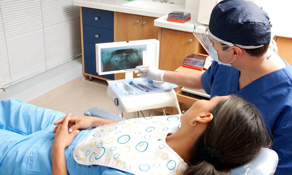 Na fotografiji je prikazana dentalna konzultacija. Pacijentica leži na stolici za zubara, prekrivena uzorkastom tkaninom, dok joj stomatolog u plavim kirurškim odorama i kapici pokazuje rendgensku snimku zuba. Lice stomatologa nije vidljivo iz razloga privatnosti. Prostorija izgleda kao dobro opremljena, moderna zubarska ordinacija s vidljivim ormarićima i raznim stomatološkim alatima. Na prozoru su rolete, što ukazuje na unutarnji prostor.