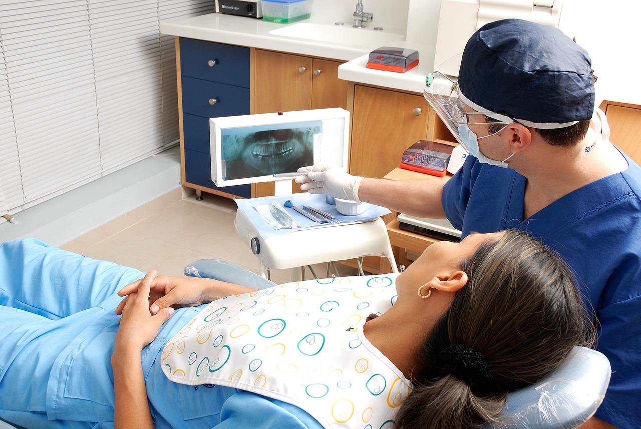 Na fotografiji je prikazana dentalna konzultacija. Pacijentica leži na stolici za zubara, prekrivena uzorkastom tkaninom, dok joj stomatolog u plavim kirurškim odorama i kapici pokazuje rendgensku snimku zuba. Lice stomatologa nije vidljivo iz razloga privatnosti. Prostorija izgleda kao dobro opremljena, moderna zubarska ordinacija s vidljivim ormarićima i raznim stomatološkim alatima. Na prozoru su rolete, što ukazuje na unutarnji prostor.