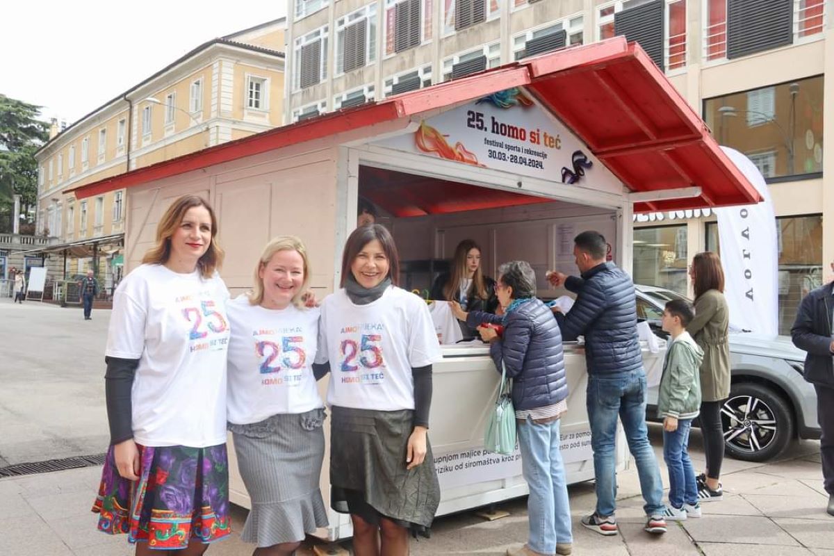 Na fotografiji je kućica za prodaju s crvenom tendom, parkirana u urbanom području s klasičnim višekatnicama u pozadini. Troje ljudi u prvom planu nosi bijele majice s oznakom “25”, a njihova lica su zamagljena zbog privatnosti. Oko prikolice se nalaze i drugi pojedinci, uključujući djecu i odrasle, koji izgledaju kao kupci ili posjetitelji. Na tendi i bočnoj strani prikolice nalazi se tekst, ali je djelomično vidljiv.