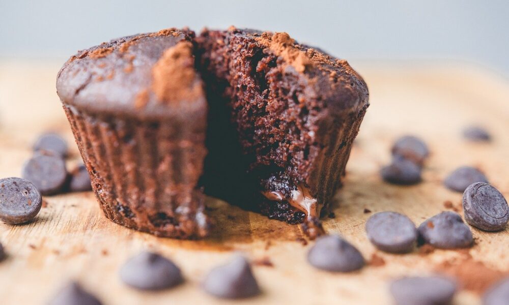 Na fotografiji je čokoladni muffin koji je prepolovljen, a oko njega su raspoređeni čokoladni komadići. Muffin izgleda sočno i ukusno, s vidljivim dijelom tekuće čokolade u sredini. Tekstura muffina je vidljivo mekana i mrvičasta, s gornjim dijelom koji ima blago hrskav sloj. Pozadina je zamagljena i neutralne boje, što usmjerava pažnju na muffin i čokoladne komadiće.