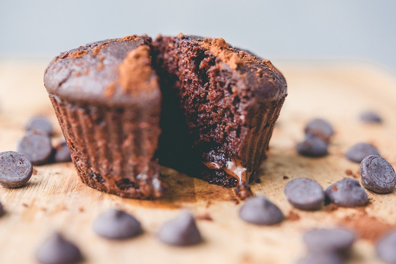 Na fotografiji je čokoladni muffin koji je prepolovljen, a oko njega su raspoređeni čokoladni komadići. Muffin izgleda sočno i ukusno, s vidljivim dijelom tekuće čokolade u sredini. Tekstura muffina je vidljivo mekana i mrvičasta, s gornjim dijelom koji ima blago hrskav sloj. Pozadina je zamagljena i neutralne boje, što usmjerava pažnju na muffin i čokoladne komadiće.