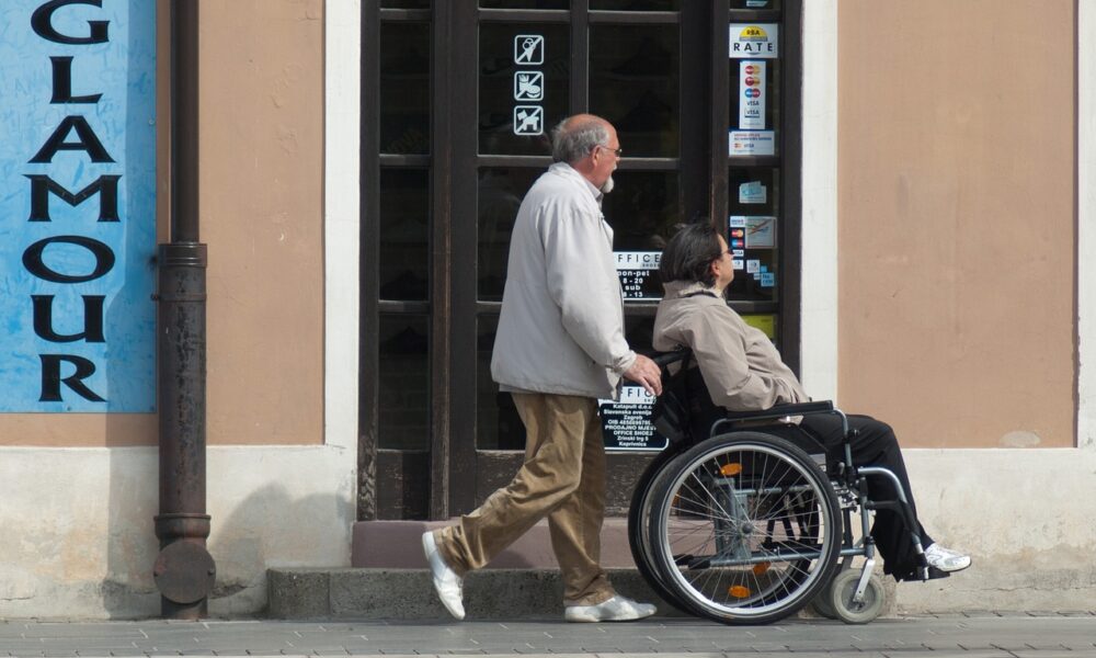 Na slici se nalaze dvije osobe; jedna hoda, a druga je u invalidskim kolicima. Prolaze pokraj trgovine s staklenim vratima na kojima se nalaze razne naljepnice. S lijeve strane slike vidljiv je veliki plavi znak s natpisom “GLAMOUR”. Zgrada ima klasičan arhitektonski dizajn s bež zidovima i smeđim obrubom oko prozora i vrata. Uz pločnik stoji metalna ulična svjetiljka obojana u crno.