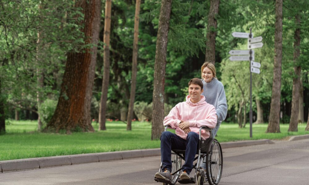 Na slici su dvije osobe, jedna je u kolicima. Žena u plavoj majici vozi u kolicima dečka u rozoj majici i crnim hlačama. Krenuli su u laganu šetnju prirodom.