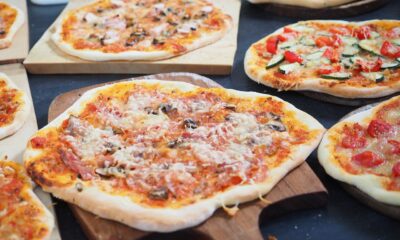 Na slici su prikazane različite vrste pizze s raznovrsnim nadjevima, poslužene na drvenim daskama. Jedna pizza u prvom planu dobro je pečena sa zlatnom korom, prelivena sirom, gljivama i moguće nekom vrstom mesa. U pozadini se nalaze druge pizze; jedna ima kriške rajčica i zelenih listova - vjerojatno bosiljak, a druga izgleda kao da ima gljive i dodatne nadjeve. Pizze su različitih oblika; neke su ovalnije dok su druge okrugle. Okruženje izgleda kao kuhinja ili stol za posluživanje gdje su pizze izložene za odabir.