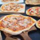 Na slici su prikazane različite vrste pizze s raznovrsnim nadjevima, poslužene na drvenim daskama. Jedna pizza u prvom planu dobro je pečena sa zlatnom korom, prelivena sirom, gljivama i moguće nekom vrstom mesa. U pozadini se nalaze druge pizze; jedna ima kriške rajčica i zelenih listova - vjerojatno bosiljak, a druga izgleda kao da ima gljive i dodatne nadjeve. Pizze su različitih oblika; neke su ovalnije dok su druge okrugle. Okruženje izgleda kao kuhinja ili stol za posluživanje gdje su pizze izložene za odabir.