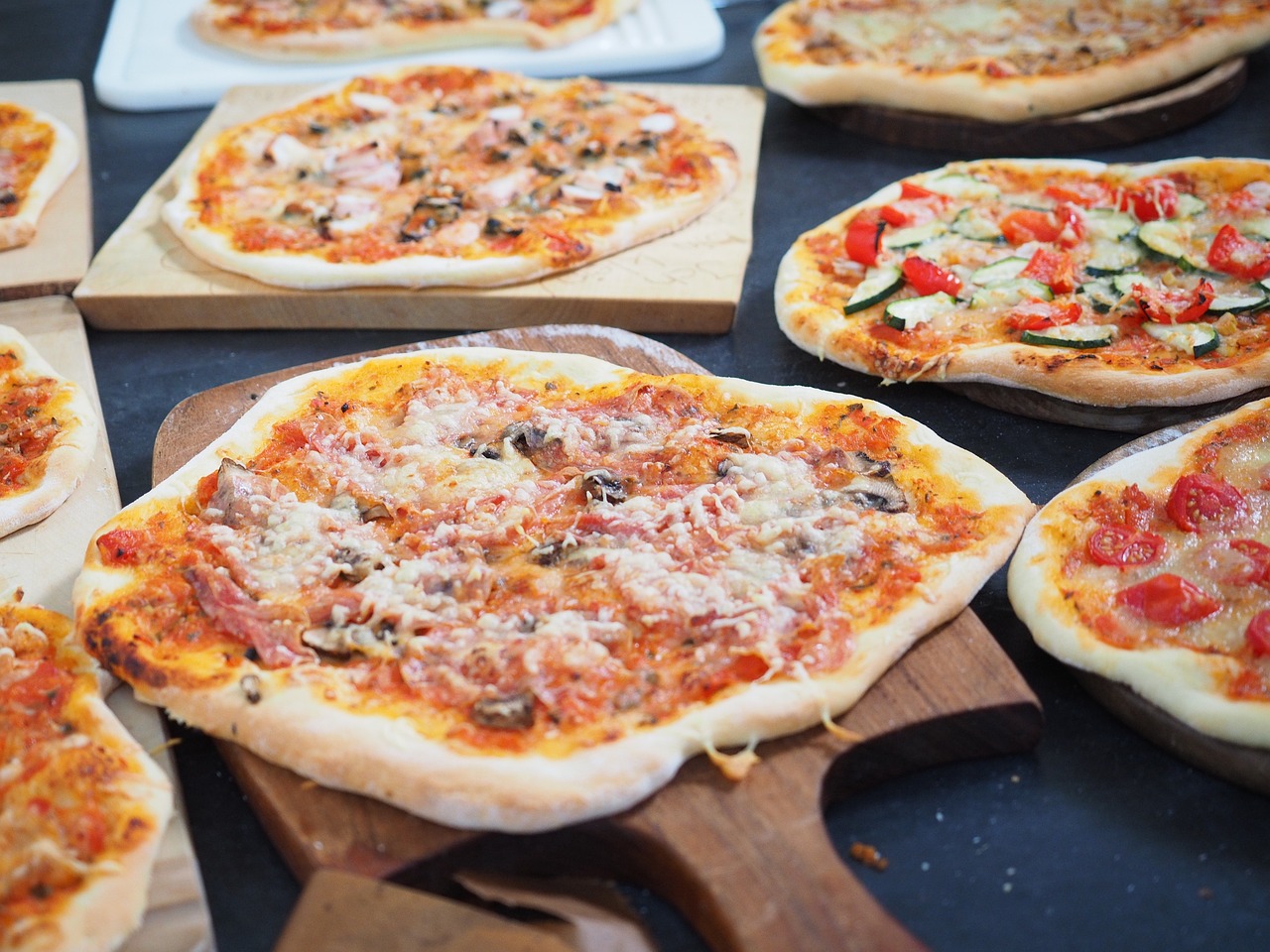 Na slici su prikazane različite vrste pizze s raznovrsnim nadjevima, poslužene na drvenim daskama. Jedna pizza u prvom planu dobro je pečena sa zlatnom korom, prelivena sirom, gljivama i moguće nekom vrstom mesa. U pozadini se nalaze druge pizze; jedna ima kriške rajčica i zelenih listova - vjerojatno bosiljak, a druga izgleda kao da ima gljive i dodatne nadjeve. Pizze su različitih oblika; neke su ovalnije dok su druge okrugle. Okruženje izgleda kao kuhinja ili stol za posluživanje gdje su pizze izložene za odabir.