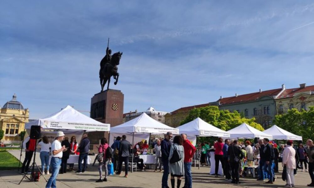 Fotografija prikazuje trg na kojem se održava neki događaj. Na trgu su postavljeni bijeli šatori, a oko njih se okupila grupa ljudi. U pozadini je spomenik konjaniku koji je postavljen na visokom postolju. Nebo je vedro s malo oblaka. Okolne zgrade s klasičnim arhitektonskim dizajnom dodaju estetsku privlačnost okruženju.