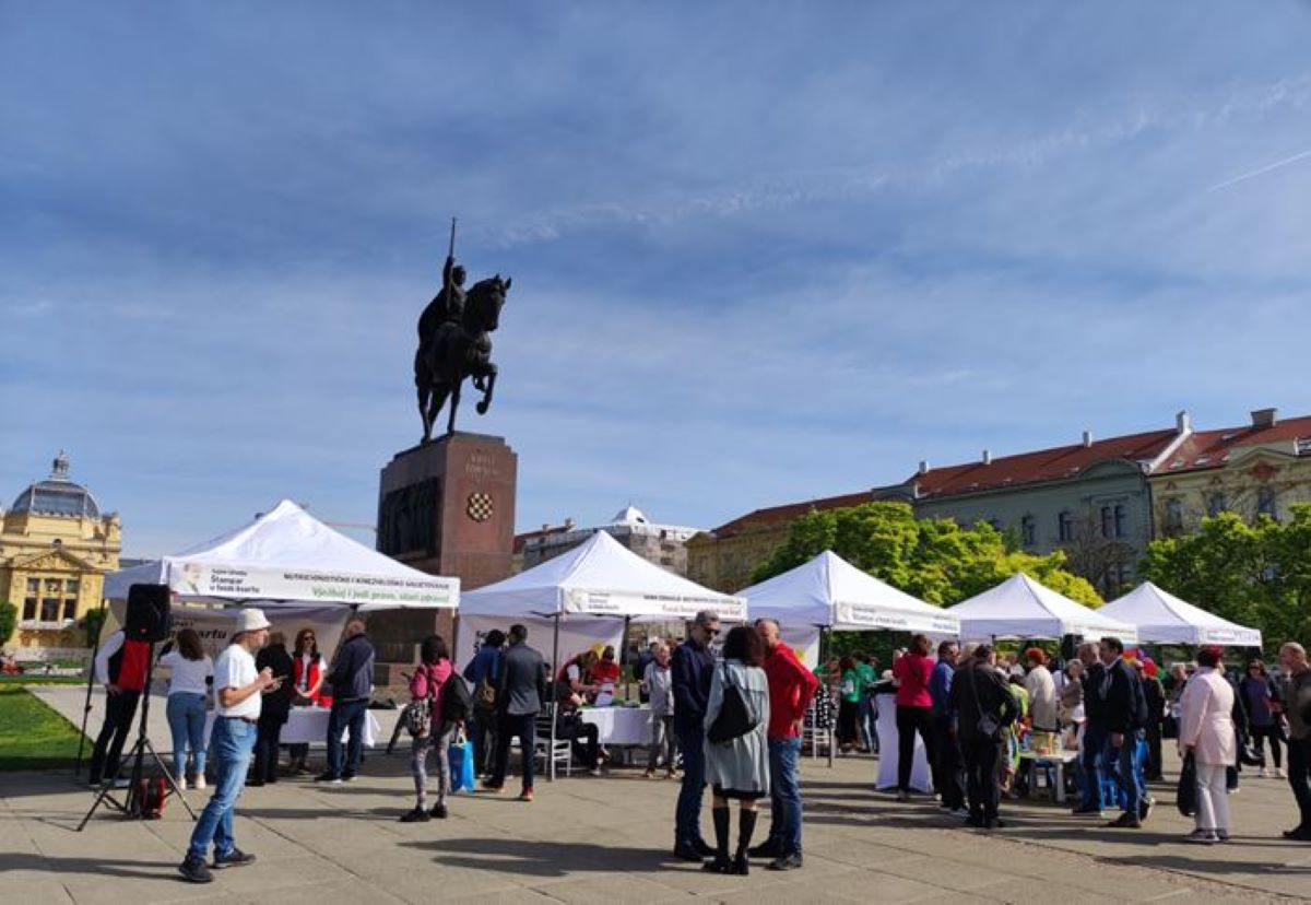 Fotografija prikazuje trg na kojem se održava neki događaj. Na trgu su postavljeni bijeli šatori, a oko njih se okupila grupa ljudi. U pozadini je spomenik konjaniku koji je postavljen na visokom postolju. Nebo je vedro s malo oblaka. Okolne zgrade s klasičnim arhitektonskim dizajnom dodaju estetsku privlačnost okruženju.