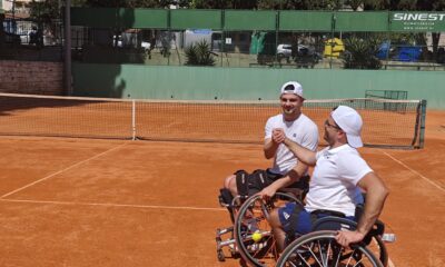 Na slici su dva tenisača u kolicima. Tenisači se pozdravljaju na teniskom terenu nakon odigrane utakmice. Jedan tenisač ima tenisku lopticu zakačenu za kolica. Oba tenisača imaju bijele majice i bijele kape. Podloga terena je zemljana.