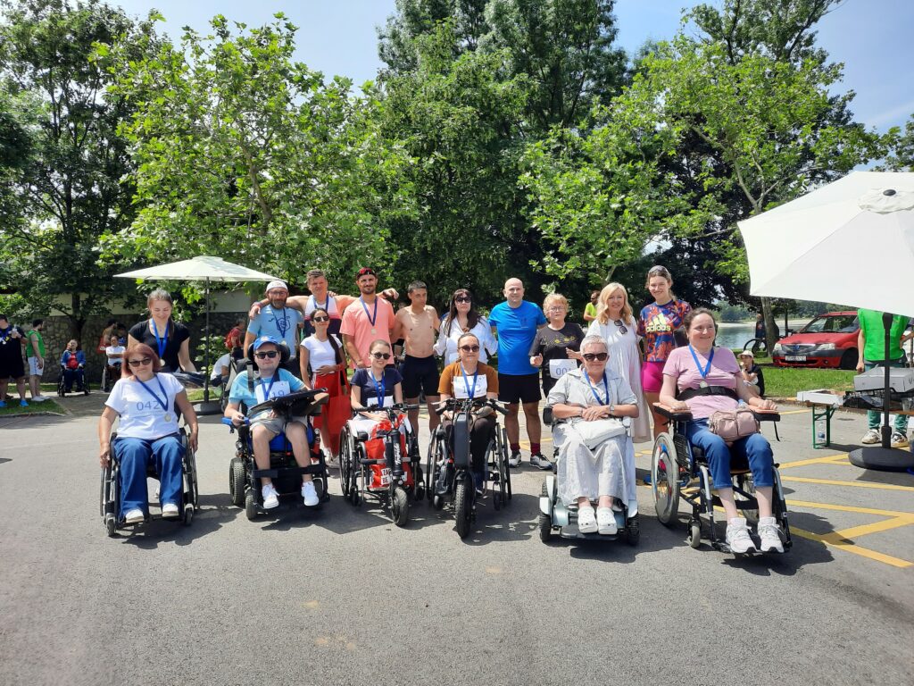 Slika prikazuje grupu osoba u invalidskim kolicima, raspoređenih u redu. Čini se da su na otvorenom, s drvećem i vedrim nebom u pozadini.