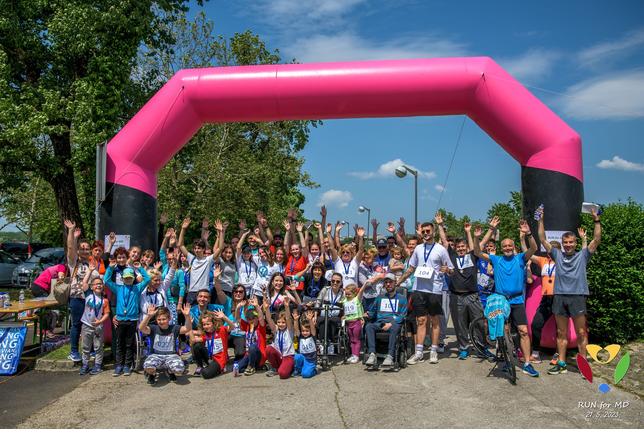 Na slici je vesela skupina ljudi koji slave ispod velikog ružičastog napuhanog balona u obliku cilja, s natpisom “RUN FOR M.D.” Vjerojatno su upravo završili neku utrku. Mnogi sudionici podižu ruke u zrak, a neki drže medalje i plakete. Nebo je plavo s nekoliko oblaka, što ukazuje na lijepo vrijeme. U pozadini se vide drveće i zelenilo. Na lijevoj strani slike nalazi se transparent, ali nije jasno što piše zbog kuta i udaljenosti.