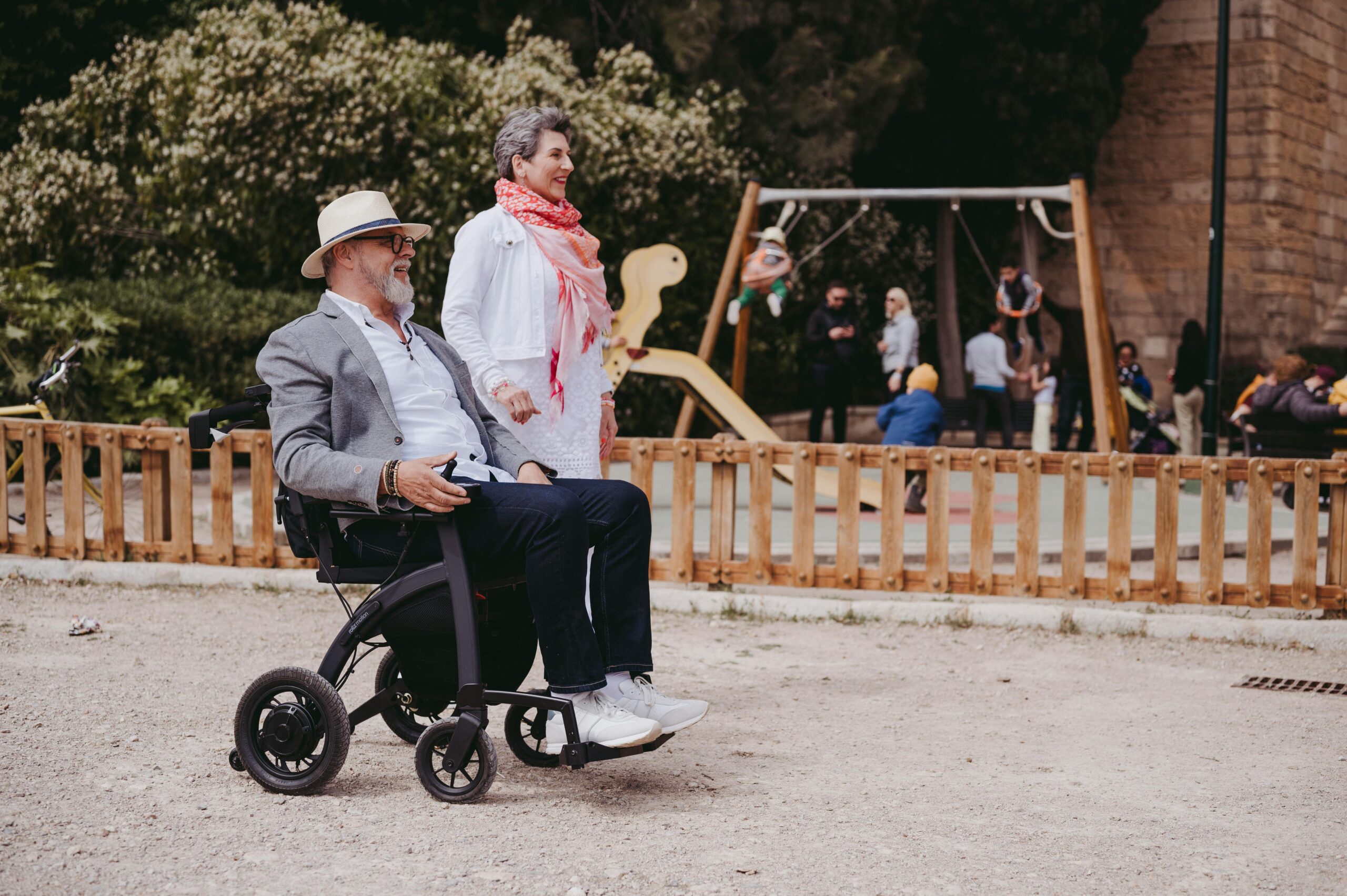 Na slici su dvije osobe: jedna sjedi u invalidskim kolicima, a druga stoji pored. Pozadina prikazuje park s dječjim igralištem gdje se ljudi okupljaju. Osoba u invalidskim kolicima nosi sivi sako, dok osoba koja stoji ima rozi šal oko vrata. Na igralištu se igraju djeca, a odrasli ih nadgledaju. Igralište je ograđeno drvenom ogradom, a oko njega su stabla koja pružaju zelenilo sceni.