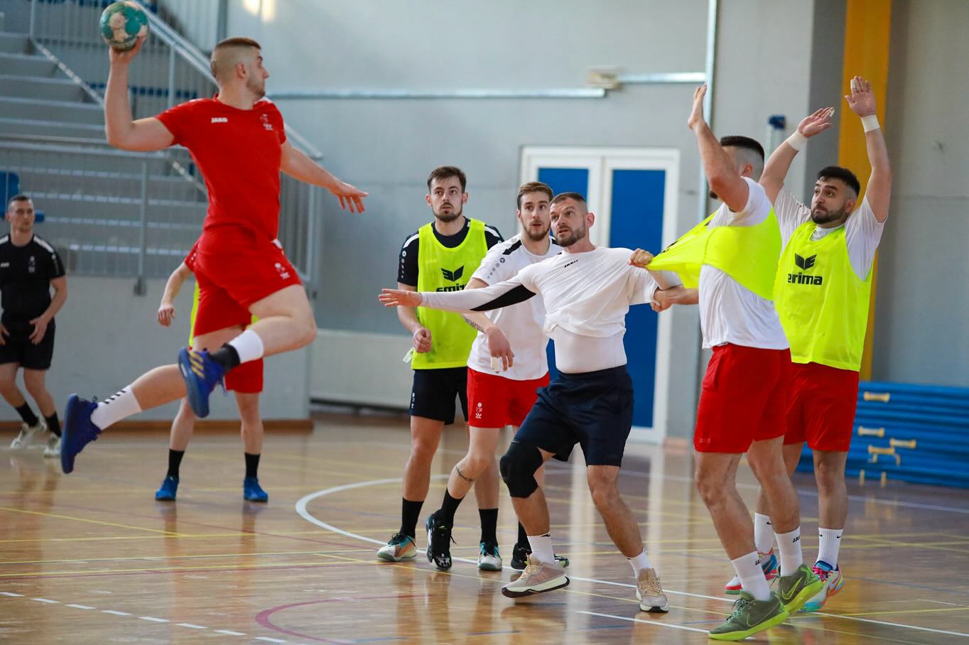 Na slici je nekoliko osoba koje igraju rukomet. Rukometaš u crvenom dresu se priprema za šut, dok ga drugi pokušavaju blokirati. Drugi igrači su obučeni u žute markere za trening, a neki nose i bijele majice.