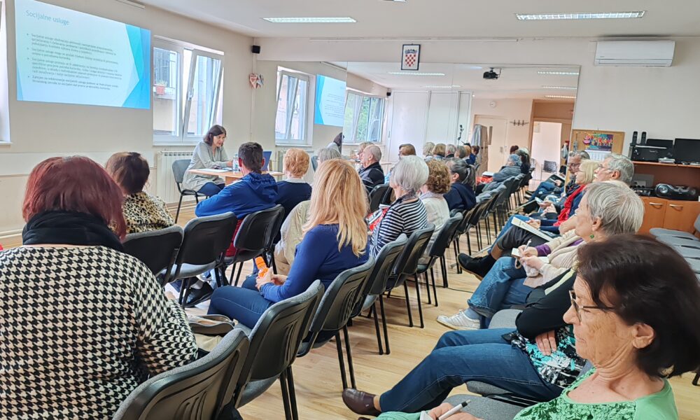 Ova slika prikazuje prostoriju s ljudima koji sjede na stolicama, okrenuti prema predavaču i projekciji na zidu. Prostorija je dobro osvijetljena prirodnim svjetlom koje dolazi kroz prozore. Na crnim stolicama sjede sudionici u više redova.