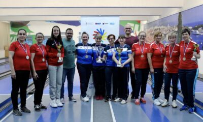 Na fotografiji je više žena, neke nose crvene, a neke plave majice, dok sve nose trenirke. Sve oko vrata imaju medalje, a neke drže i pehare. Fotografija je slikana u kuglani.