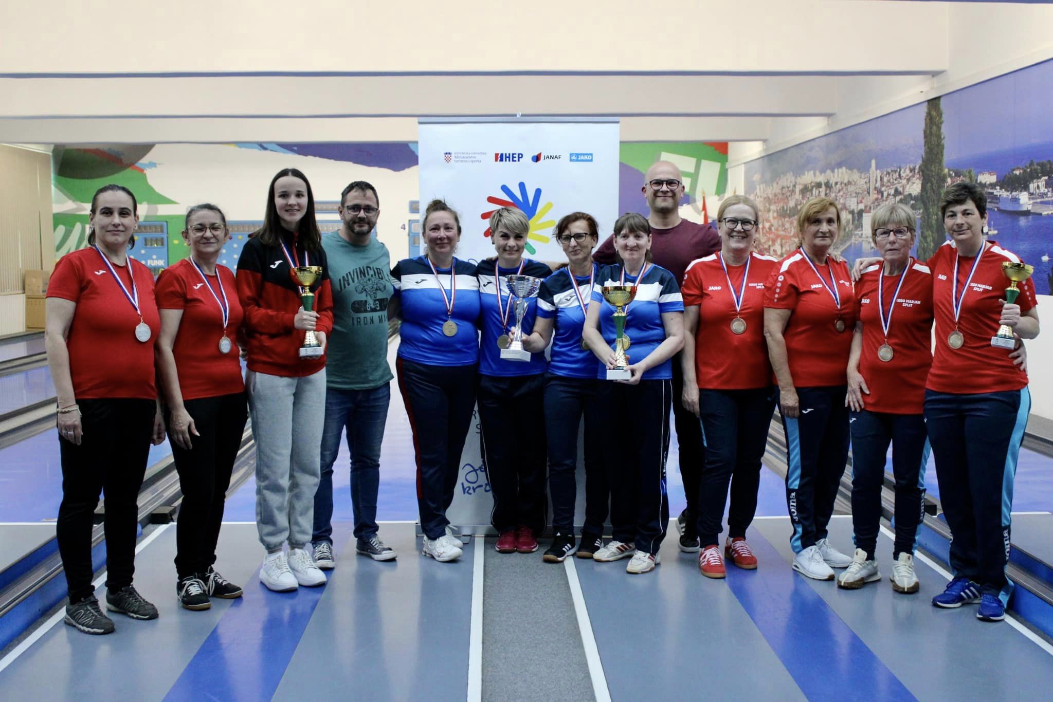 Na fotografiji je više žena, neke nose crvene, a neke plave majice, dok sve nose trenirke. Sve oko vrata imaju medalje, a neke drže i pehare. Fotografija je slikana u kuglani.