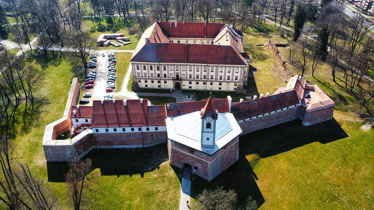 Ova slika prikazuje zračni pogled na velku, pravokutnu tvrđavu s crvenim krovovima okruženu obrambenim zidovima. Na jednom od uglova tvrđave ističe se bijeli stražarni toranj. Tvrđava je smještena u zelenom području s drvećem, a uz nju se nalazi parkiralište.