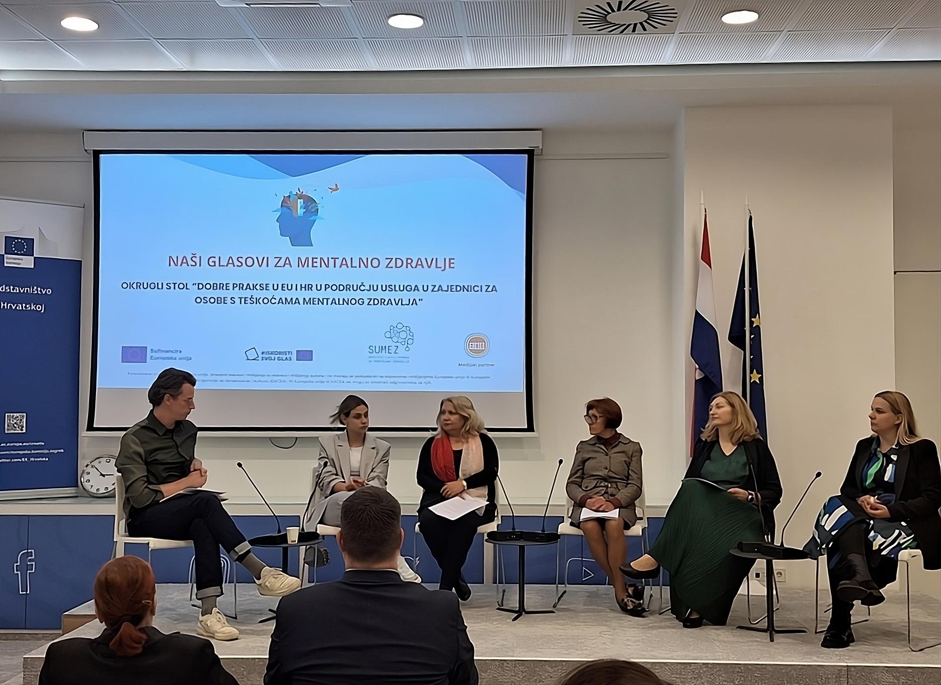 Ova slika prikazuje panel diskusiju s pet osoba koje sjede na stolicama ispred plavog zida s projekcijskim ekranom. Na ekranu je prezentacija, a sudionici se čini da su uključeni u raspravu ili prezentaciju. Desno od njih su zastave, uključujući zastavu Europske unije i zastavu Hrvatske.