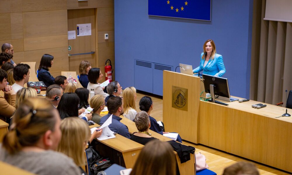 Slika prikazuje predavaonicu u kojoj osoba drži predavanje. Publika pažljivo sluša, a neki čak i bilježe. Na zidu iza govornice vidi se zastava Europske unije.