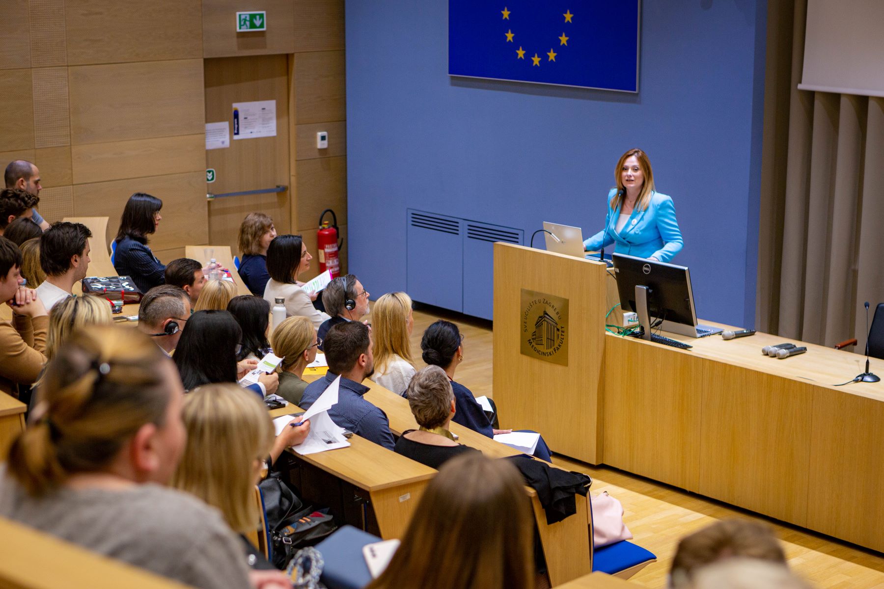 Slika prikazuje predavaonicu u kojoj osoba drži predavanje. Publika pažljivo sluša, a neki čak i bilježe. Na zidu iza govornice vidi se zastava Europske unije.