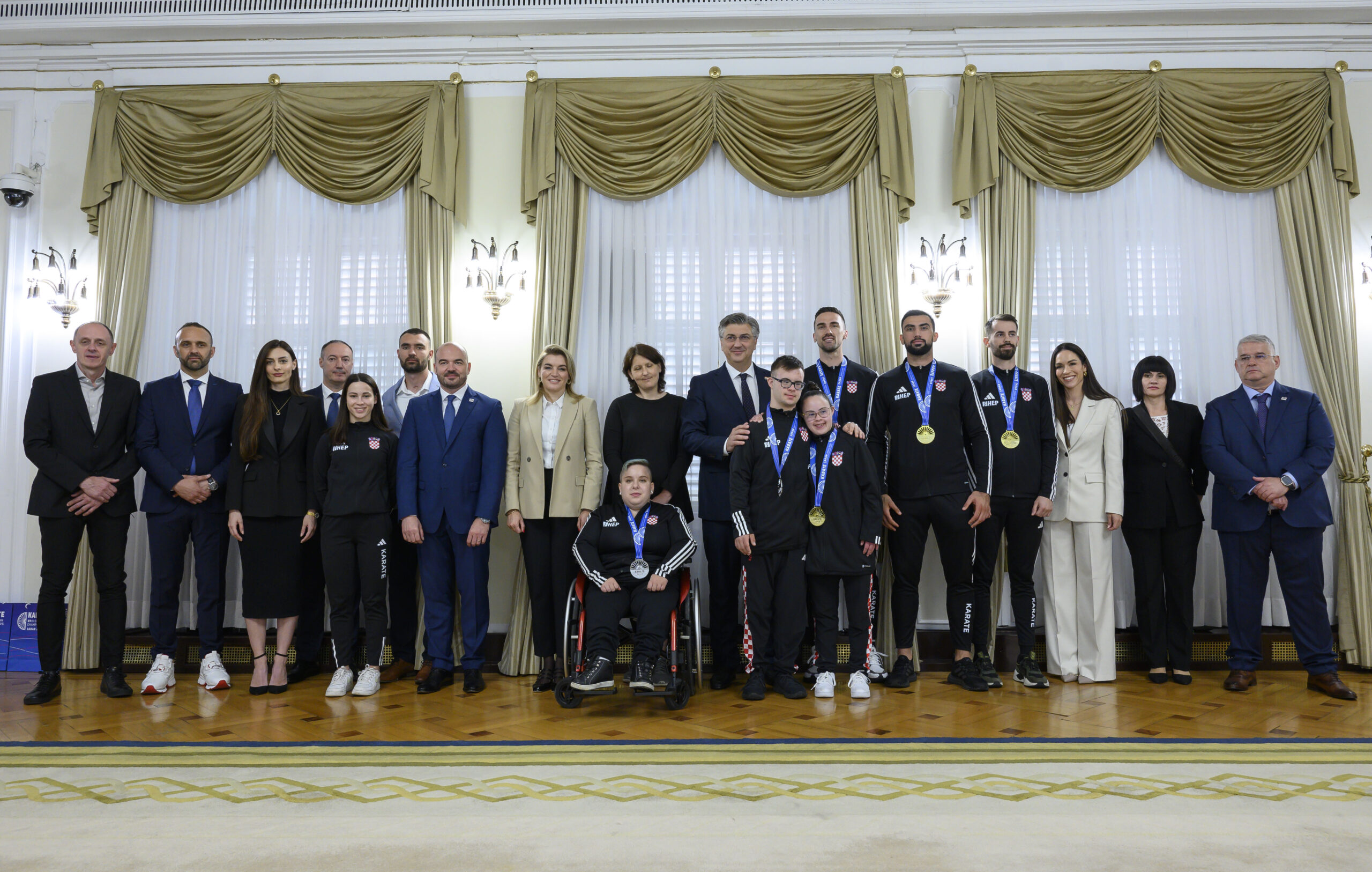 Na slici je više osoba, jedna osoba je u invalidskim kolicima, a druge stoje. Većina ih nosi medalje oko vrata Par je ljudi u odjelima dok su ostali u trenirkama.