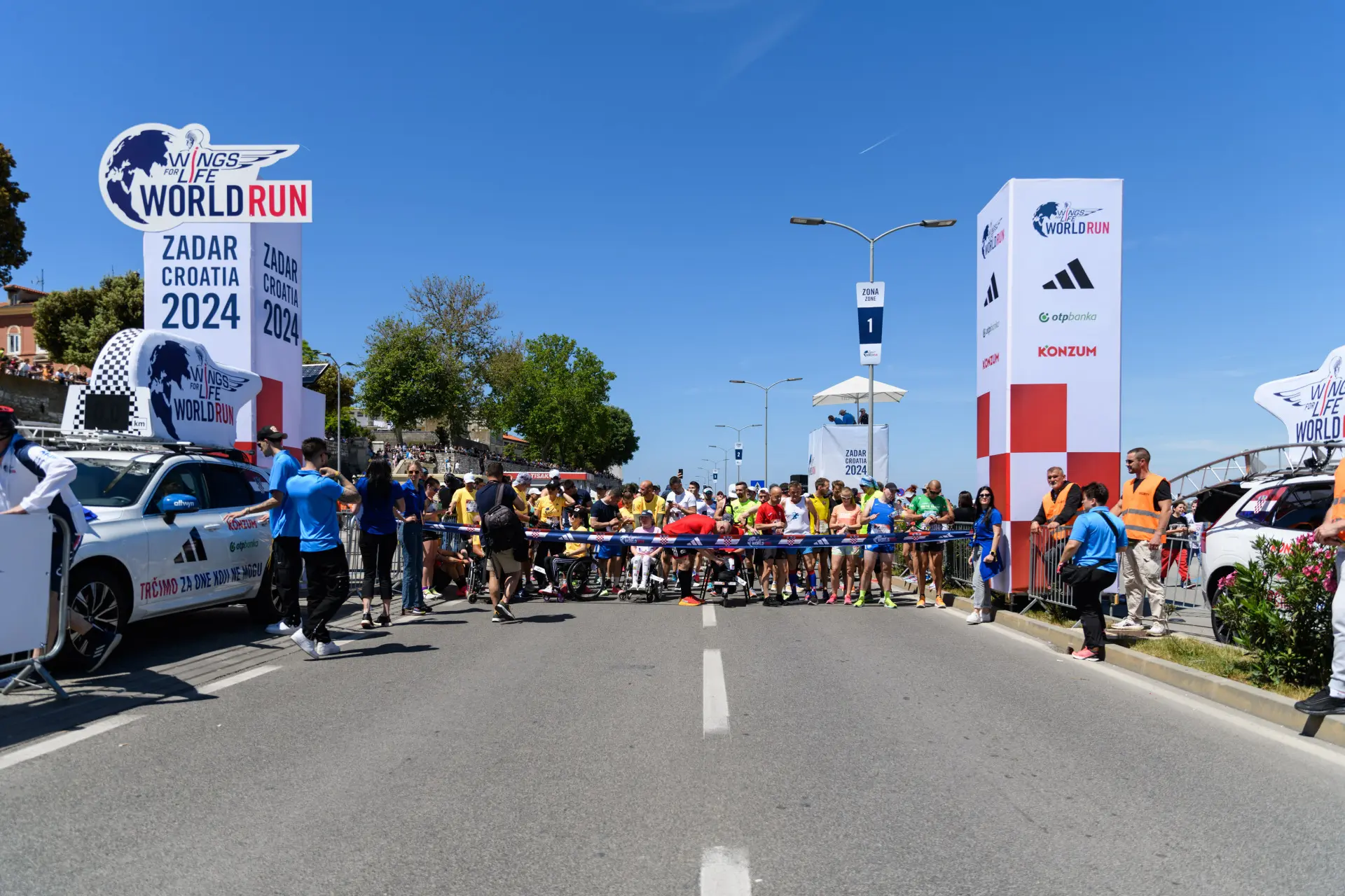 Na slici su trkači koji se pripremaju za utrku. Na jednoj strani piše Zadar Croatia Wings for life i parkiran je automobil na kojem isto piše. S druge strane se vide sponzori utrke, kao što su Adidas i Konzum te također piše World Run. Vide se i crveno bijele kockice.