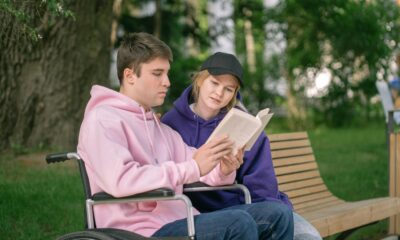 Na slici su dvoje ljudi. Jedna osoba je u kolicima i čita knjigu, a druga sjedi pored njega. Nalaze se u ugodnom ambijentu okruženi prirodom.