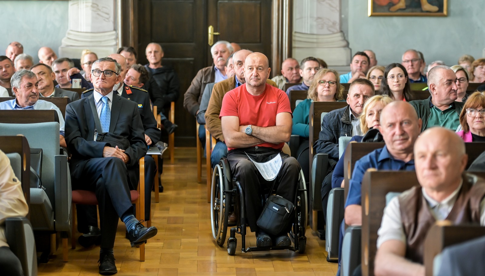 Slika prikazuje grupu ljudi koji sjede u prostoriji s visokim stropom i umjetničkim slikama na zidovima. Prostorija je dobro osvijetljena, što može ukazivati na događaj ili okupljanje. Osoba u prvom planu sjedi u invalidskim kolicima, nosi crvenu majicu i ima papire u krilu.