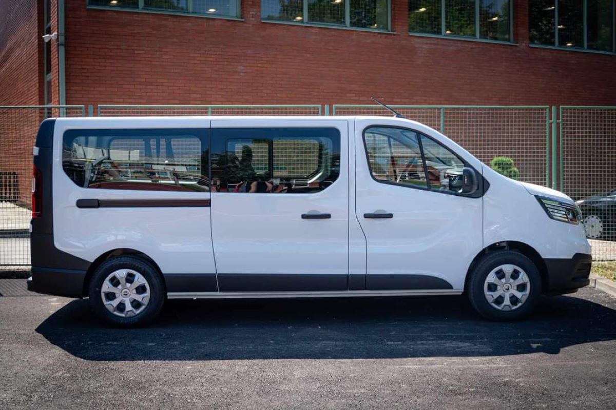 Na fotografiji je bijeli kombi parkiran ispred crvene zgrade. Kombi ima tamne prozore na stražnjem dijelu i odražava se okolina.