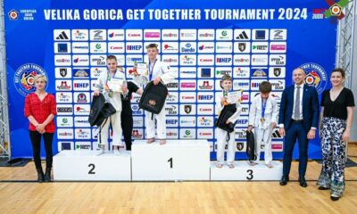 Na slici je nekoliko ljudi. četvero ih nosi kimono, dok je troje obučeno u običnu robu. Na zidu je veliki plavi plakat na kojem piše ‘Velika Gorica Get Together Tournament 2024’, na plakatu su oznake sponzora natjecanja.