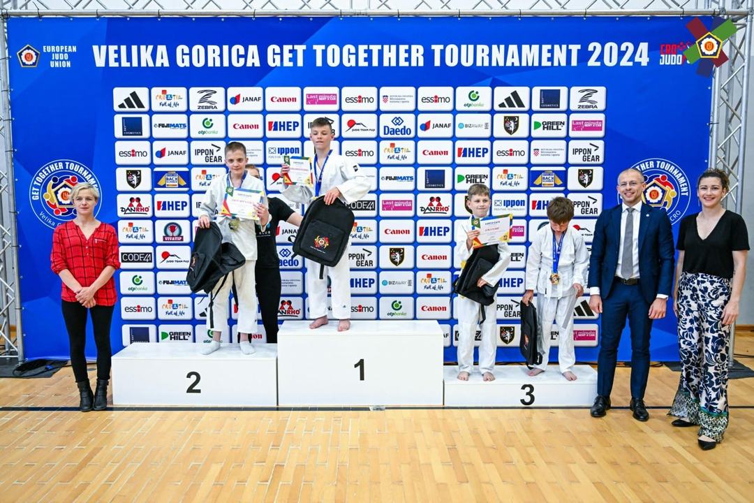 Na slici je nekoliko ljudi. četvero ih nosi kimono, dok je troje obučeno u običnu robu. Na zidu je veliki plavi plakat na kojem piše ‘Velika Gorica Get Together Tournament 2024’, na plakatu su oznake sponzora natjecanja.