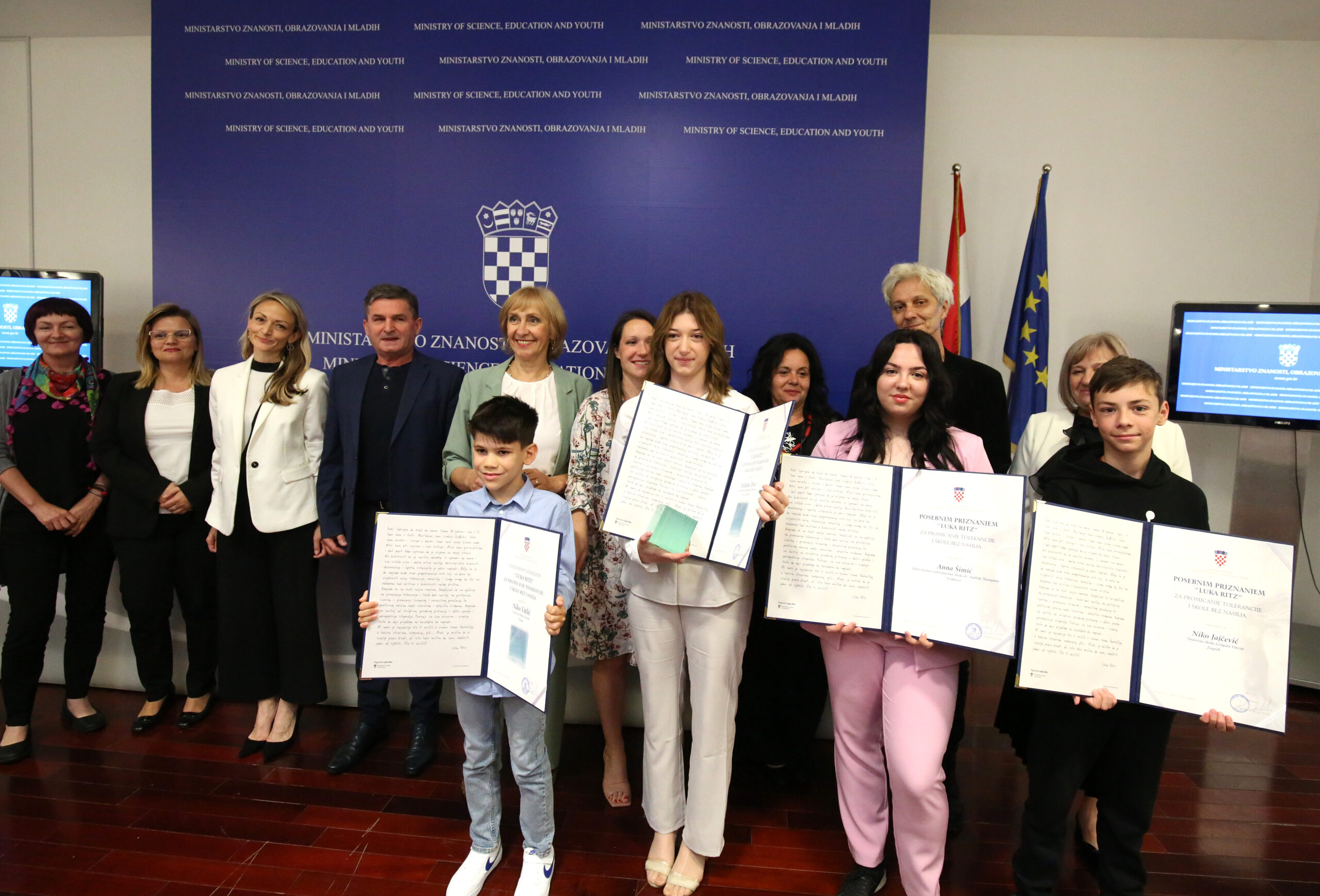 Na slici je grupa ljudi koja stoji u redu ispred plavog zida s natpisom "MINISTARSTVO ZNANOSTI, OBRAZOVANJA I MLADIH". Ispred njih su djeca koja drže velike certifikate i priznanja. Svi su nasmiješeni i poziraju za kameru. Na zidu su također zastave Hrvatske i Europske unije.