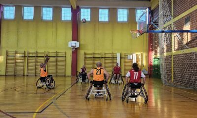 Na slici su ljudi u invalidskim kolicima koji igraju košarkašku utakmicu.. Jedna ekipa je u narančastim, a druga u crvenim dresovima, Igrač u narančastom dresu uputio je udarac prema košu. Zidovi dvorane su žute boje,, dok je parket smeđe boje, a na vrhu dvorane nalazi se više malih prozora.