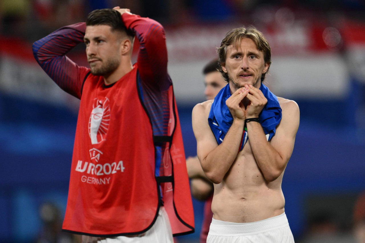 Na slici su prikazana dva nogometaša u emotivnom trenutku. Jedan od njih nosi crveni marker s natpisom “EURO 2024 GERMANY” i izgleda razočarano držeći ruke na glavi. Drugi nogometaš je bez majice, pokriva lice rukama i čini se uznemiren. Ova slika može odražavati snažne emocije nakon važne nogometne utakmice.