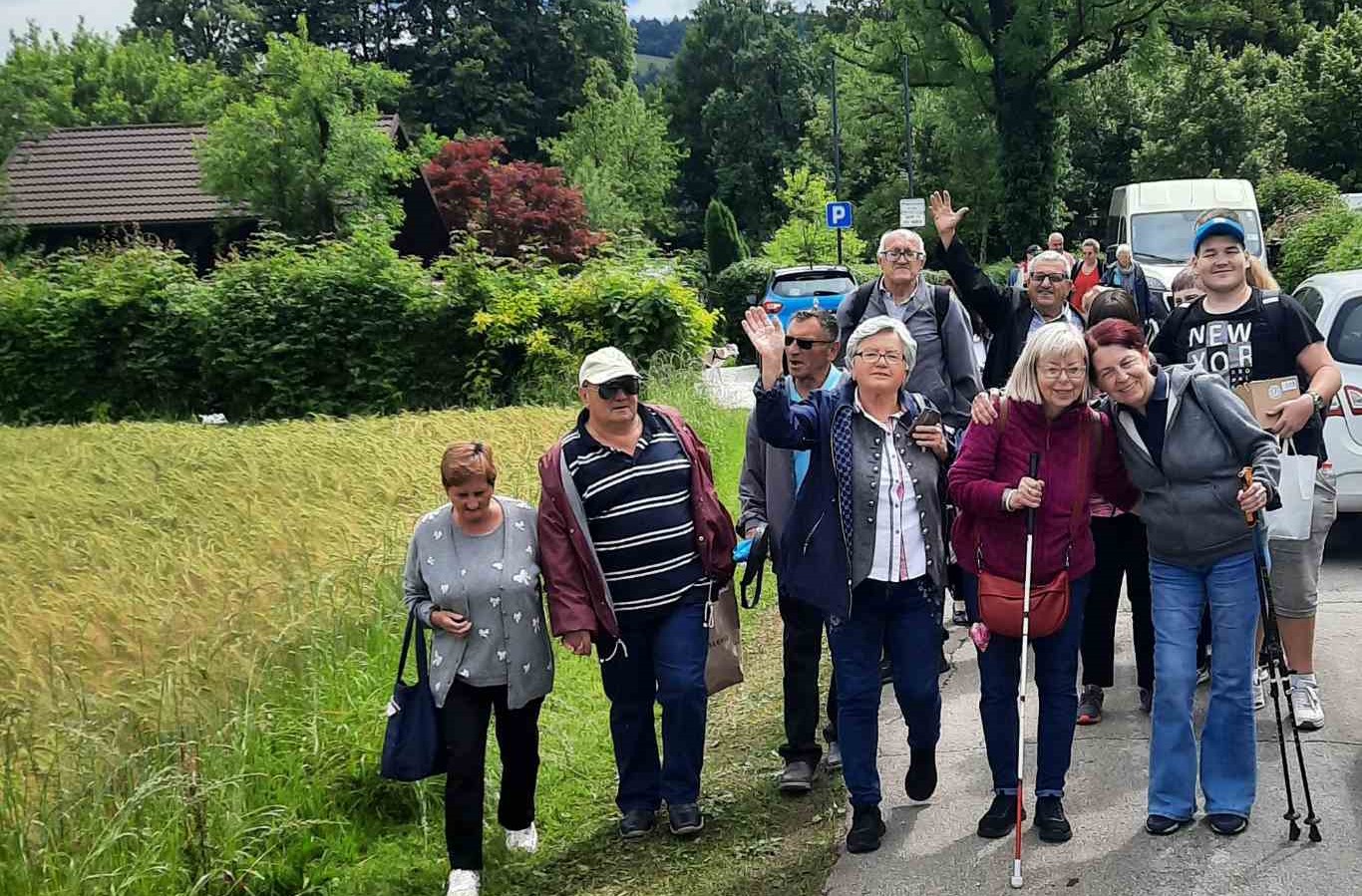 Na slici se nalazi grupa ljudi na otvorenom, s lica koja su zamagljena radi privatnosti. Čini se da su na stazi ili cesti okruženi zelenilom i drvećem. Neki od njih drže štapove za hodanje, što sugerira da bi mogli biti na pješačenju ili šetnji. Postoji znak s oznakom ‘P’ koji ukazuje na parkiralište, a u pozadini su vidljiva vozila. Okruženje izgleda ruralno ili polururalno.