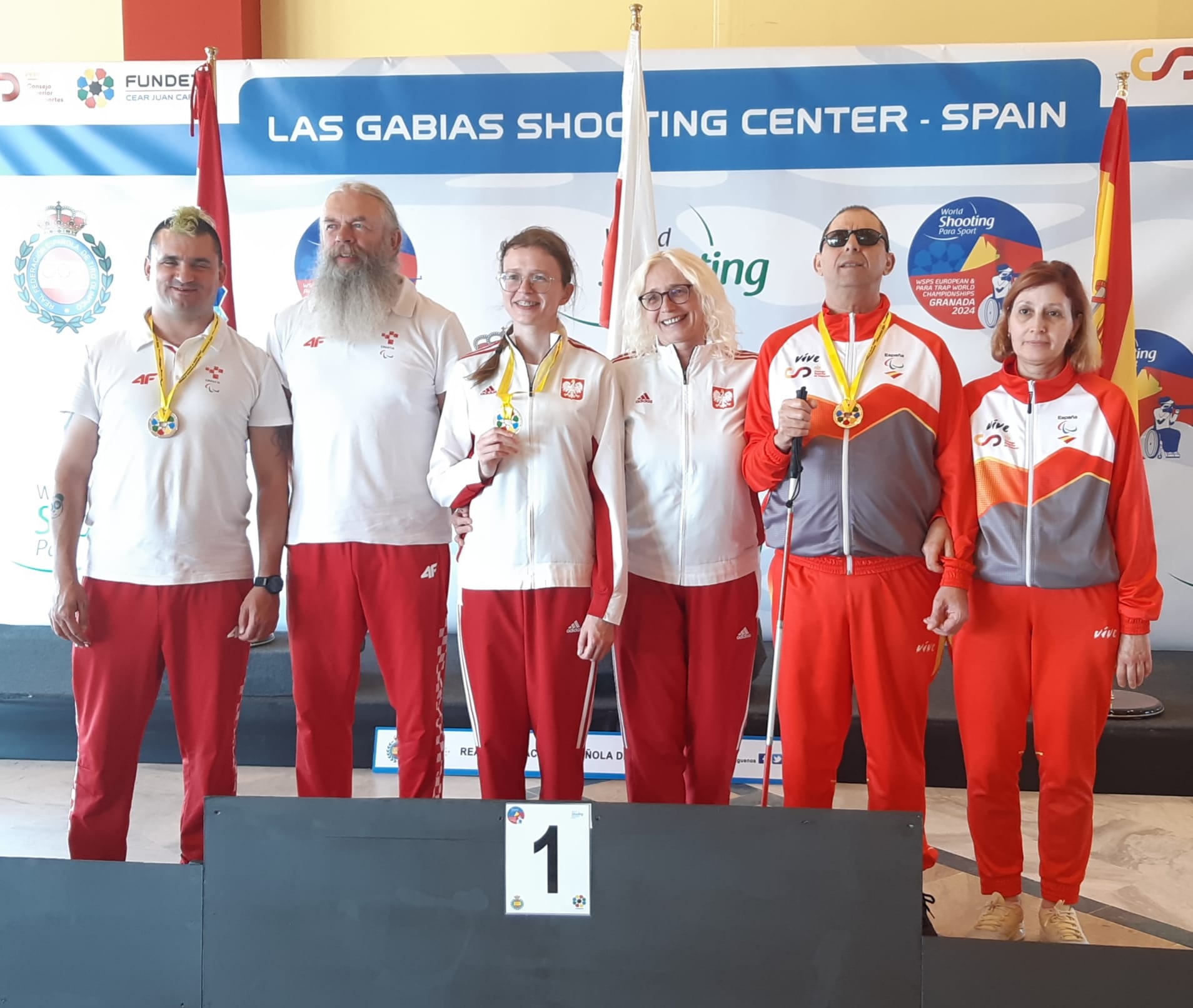 Na slici je grupa od šest osoba koje stoje na pobjedničkom postolju. Svi nose sportske uniforme i medalje oko vrata. Troje osoba s lijeve strane su u bijelim uniformama, a troje s desne strane u crvenim. U pozadini se vide zastave i transparent na kojem piše “LAS GABIAS SHOOTING CENTER - SPAIN”. Radi se o natjecanju u streljaštvu, što se može zaključiti po tekstu napisanom na transparentu.