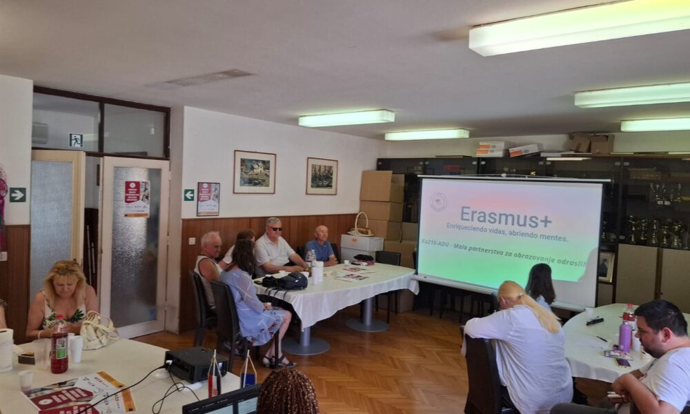 Sastanak u okviru projekta Erasmus+ gdje sudionici sjede za stolovima i slušaju prezentaciju na kojoj piše "Erasmus+".