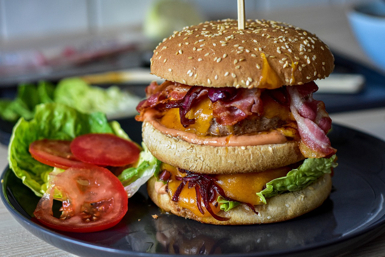 Na fotografiji je prikazan dvostruki hamburger koji sadrži dvije pljeskavice, sir, slaninu i umak. Hamburger je poslužen na tanjuru s prilogom od svježe salate i kriški rajčice. Ovaj obrok bi mogao privući ljubitelje brze hrane i može se koristiti u marketinške svrhe za restorane ili recepte.