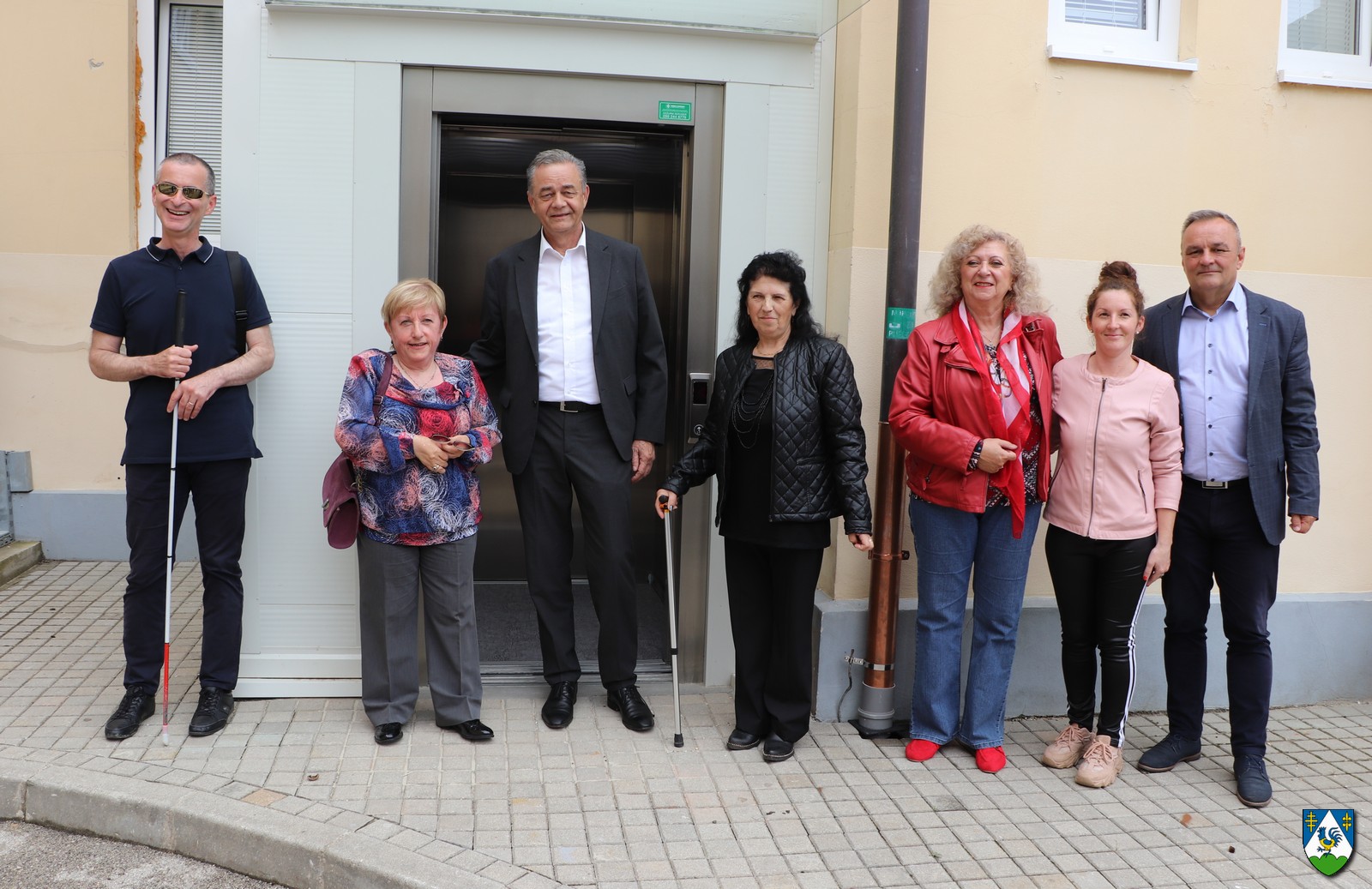 Na fotografiji je grupa od osam osoba koje stoje ispred ulaza u zgradu. Neki su formalno odjeveni u odijela, dok su drugi u ležernoj odjeći. Zgrada iza njih je moderna, siva boje s metalnim okvirima na prozorima i vratima. Postoji znak koji bi mogao biti službeni grb ili logo, ali nije jasno vidljiv.