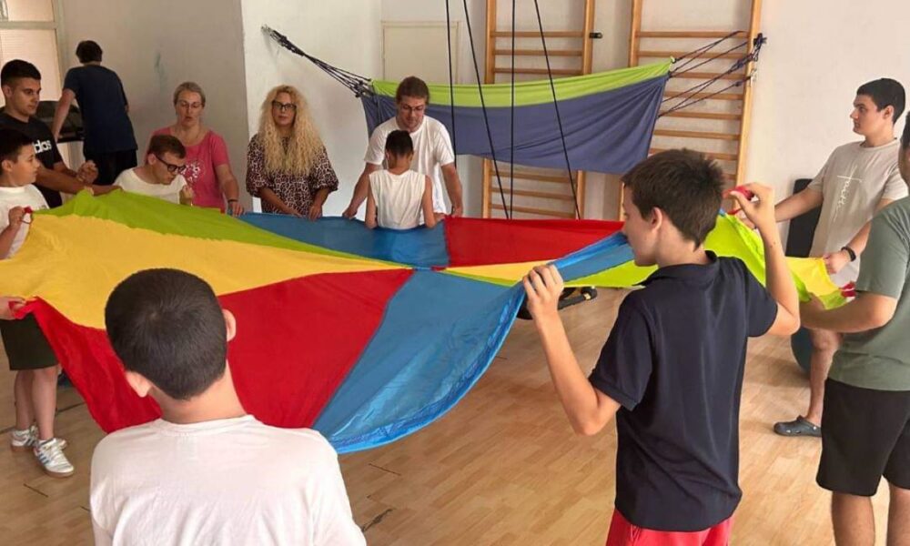 Na fotografiji je skupina djece koja se igra s velikim šarenim padobranom u tjelesnoj dvorani. Djeca su okrenuta leđima prema kameri, a lica nekih su zamagljena. U pozadini se vide gimnastičke sprave, a pod je parketiran. U donjem desnom kutu je logo koji podsjeća na grb ili pečat. Slika odražava suradnju i fizičku aktivnost, što je važno za razvoj djece.