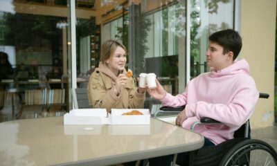 Na slici je dvoje ljudi koji piju kavu iz plastičnih bijelih čaša. Muškarac je u kolicima, a žena sjedi pokraj njega. Krafne su također na stolu, a žena je pojela pola jedne.