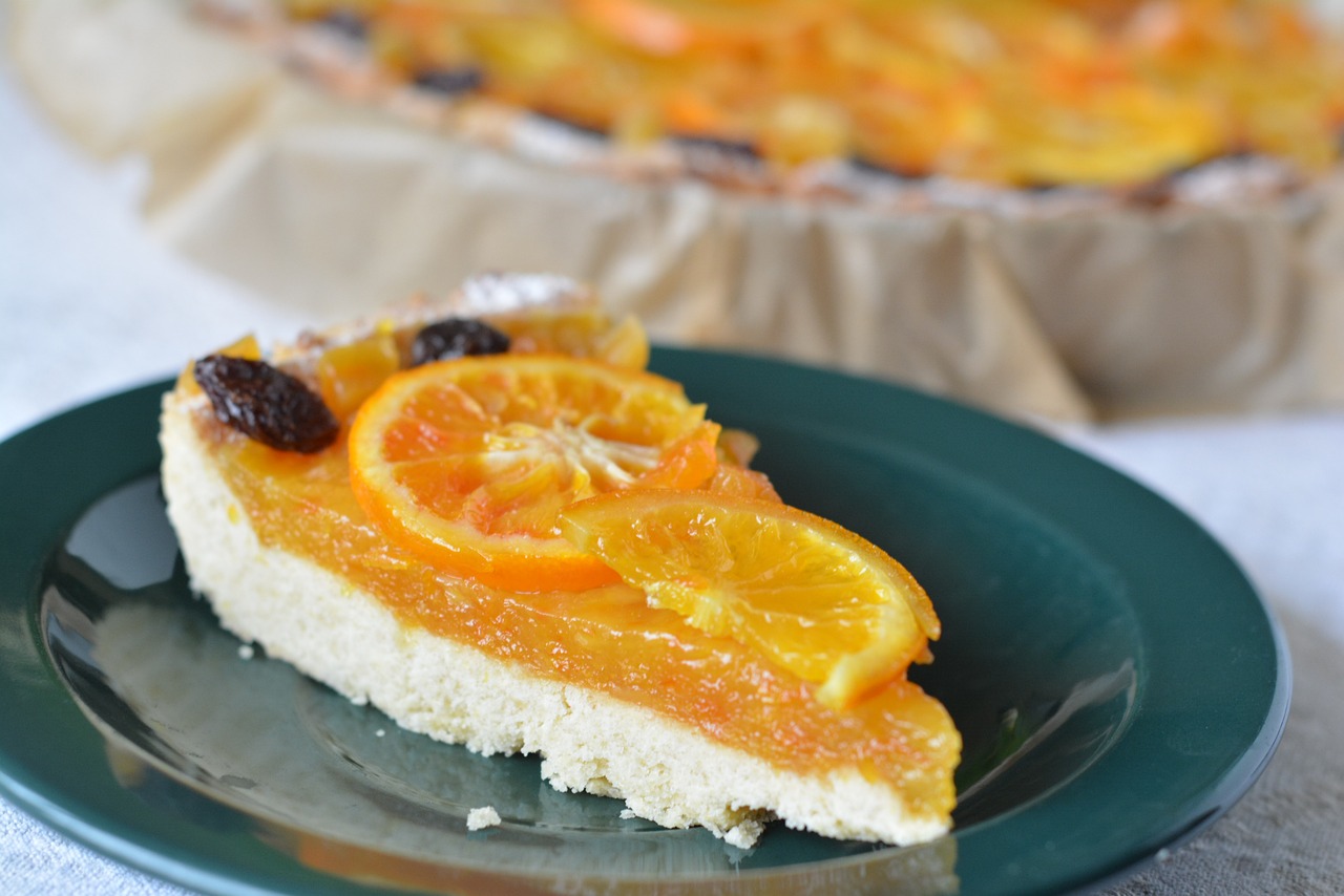 Na fotografiji je prikazan komad torte s narančama na zelenom tanjuru. Torta izgleda kao cheesecake ili neka vrsta kolača s kremastom teksturkom, prekrivena slojem svježe narezanih naranči koje su posložene u krug. Boje su živopisne i kontrast između narančaste boje voća i zelene boje tanjura čini sliku privlačnom. Pozadina je nejasna, ali se može naslutiti da je torta postavljena na stol prekriven smeđim papirom ili tkaninom. Fotografija bi mogla biti zanimljiva zbog prikaza apetitlih desertnih jela koja mogu privući pažnju ljubitelja slatkiša ili onih koji traže inspiraciju za recepte.