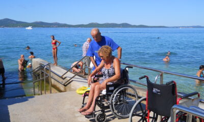Na slici su dvoje ljudi na plaži. Žena je u invalidskim kolicima, a muškarac u plavoj majici joj pomaže, Na slici je još nekoliko ljudi u kupaćim kostimima koji uživaju na plaži.