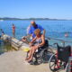 Na slici su dvoje ljudi na plaži. Žena je u invalidskim kolicima, a muškarac u plavoj majici joj pomaže, Na slici je još nekoliko ljudi u kupaćim kostimima koji uživaju na plaži.