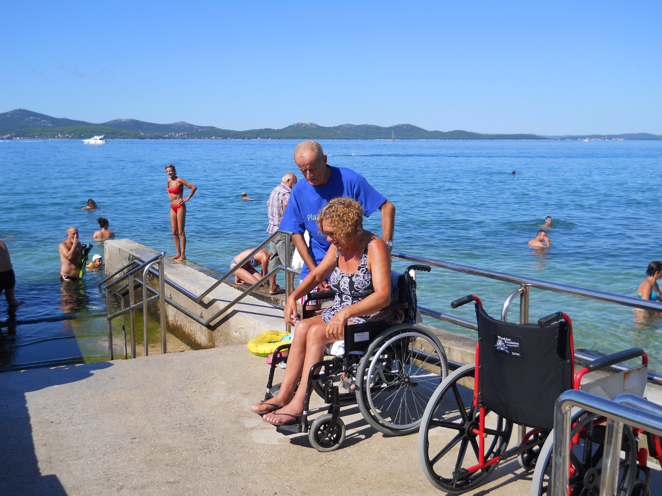 Na slici su dvoje ljudi na plaži. Žena je u invalidskim kolicima, a muškarac u plavoj majici joj pomaže, Na slici je još nekoliko ljudi u kupaćim kostimima koji uživaju na plaži.