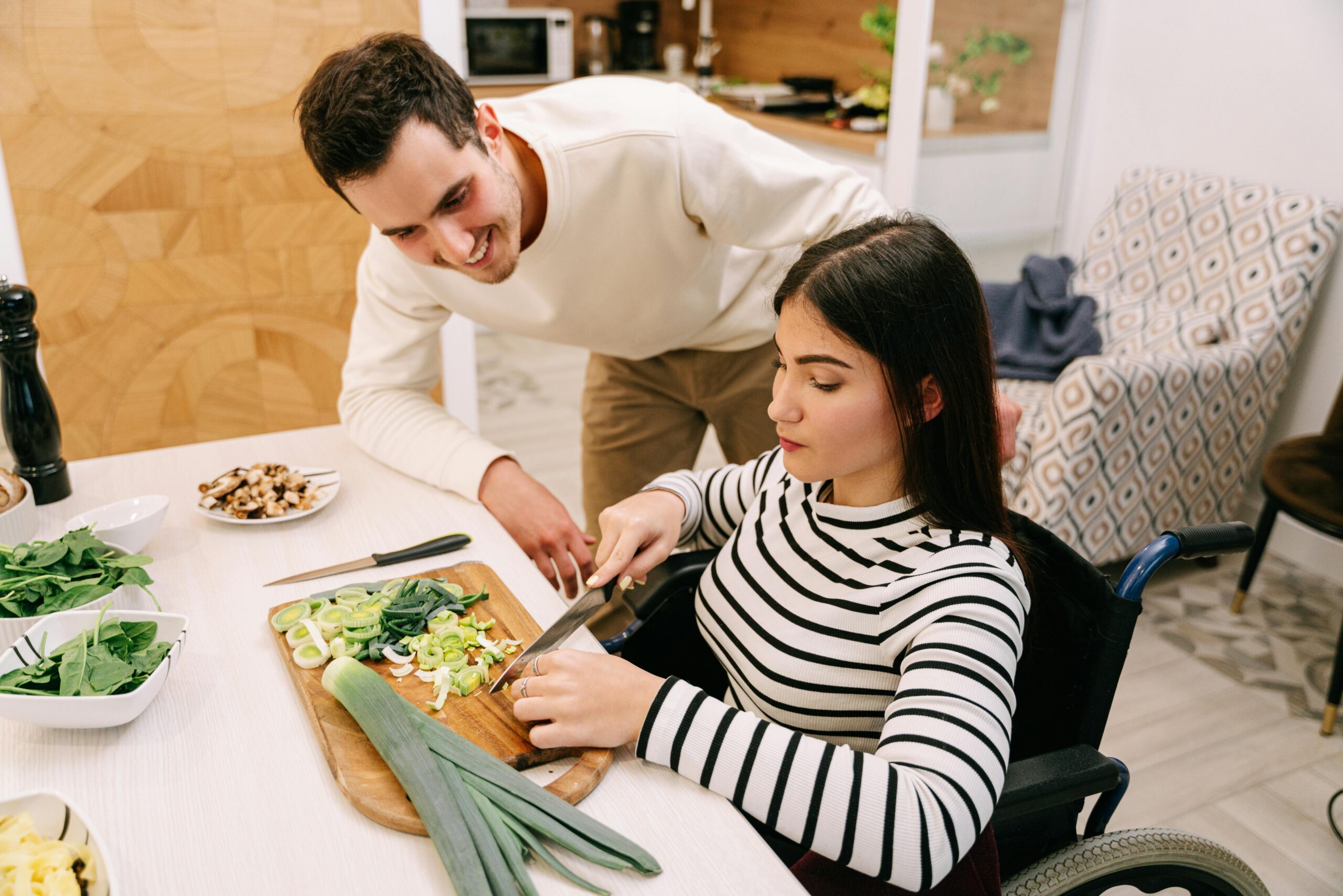 Na slici su dvije osobe koje pripremaju hranu u kuhinji. Osoba u invalidskim kolicima reže povrće na drvenoj dasci, dok je druga promatra. Na stolu se nalazi različito povrće i posude, što ukazuje na to da se priprema obrok.